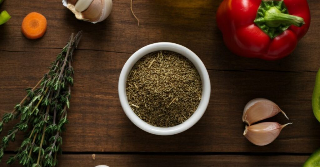 A close-up, flat-lay shot of a rustic wooden table with a small bowl of dried thyme surrounded by fresh thyme sprigs, olive oil, garlic cloves, and a few colorful vegetables like carrots and bell