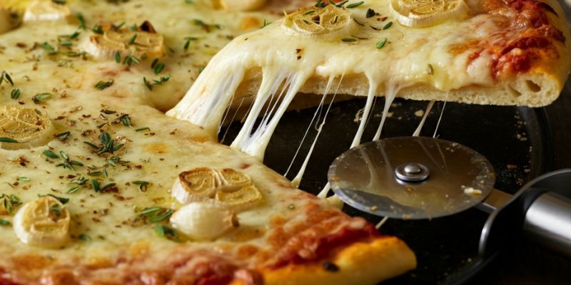A freshly baked pizza with melted cheese, roasted garlic, and thyme sprinkled on top, with a pizza cutter and a slice being pulled away