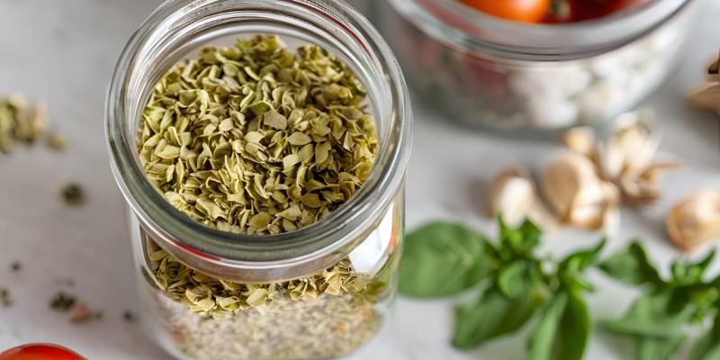 A glass jar of dried oregano placed on a kitchen counter with fresh tomatoes and garlic in the background.